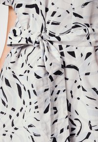 Vit tyg med abstrakta svarta och grå oroligheter, med en knuten bältesdetalj, slät textur och avslappnad silhuett.