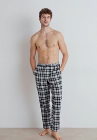 Pantalons à carreaux noir et blanc, coupe loose, avec poches latérales et taille élastique, portés par un modèle sans chemise, debout pieds nus.