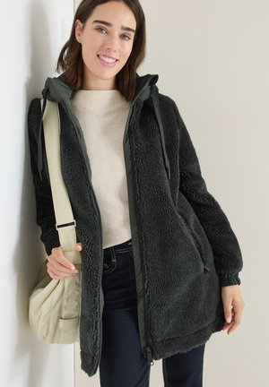 Veste en polaire gris foncé avec capuche, présentant une texture douce et des poignets élastiques. Associée à un sac à main beige clair et un pull crème.