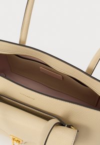 Beige leren handtas met een gladde textuur, gouden hardware en donkere randafwerking. Voorzien van een ritssluiting en een ruime binnenkant met een opbergvak.