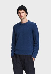 Maglione lavorato a maglia blu navy con scollatura rotonda e polsini a costine. Indossato con pantaloni neri, che presentano una finitura testurizzata e una vestibilità morbida.
