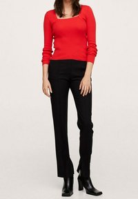 Donna in un top rosso a maniche lunghe a coste, pantaloni neri dritti con fessure frontali e stivaletti neri con tacco, in piedi su uno sfondo bianco.