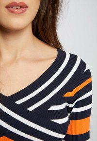 Maglione a righe blu navy e bianche con scollo a V profondo, caratterizzato da dettagli arancioni e texture a coste. Primo piano del colletto e delle spalle.