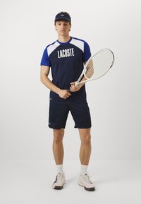 Navy Lacoste atletska majica z belimi detajli, v kombinaciji z mornarskimi kratkimi hlačami. Model drži teniško lopar, nosi ustrezen ščitnik za oči in športne čevlje.