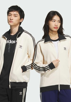 Dva pojedinca nose kremaste rebraste jakne na patentni zatvarač s crnim naglascima i tri crne pruge na rukavima, uparene s crno-bijelim gornjim dijelovima.