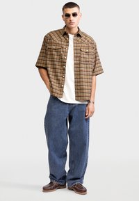 Een bruin en geel geruit kortemouwen overhemd, een witte T-shirt met ronde hals eronder, relaxed-fit blauwe jeans en bruine schoenen met een gestructureerde zool.
