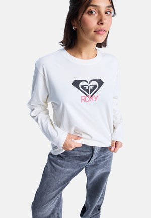 White long-sleeve t-shirt with a black and pink heart logo, made of soft cotton fabric. Model wears gray denim pants.