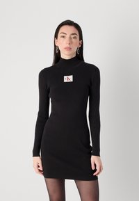 Model pozira u uskoj crnoj haljini s rebrastim uzorkom, visokim ovratnikom i oznakom logotipa, uz dodatak naušnica na prstenove, ispred jednostavne pozadine.