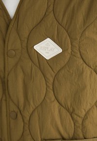 Vesta matlasată în verde oliv, cu un patch în formă de diamant de culoare albă cu inscripția "REPLAY 1981". Cusături rotunjite și două butoane vizibile.