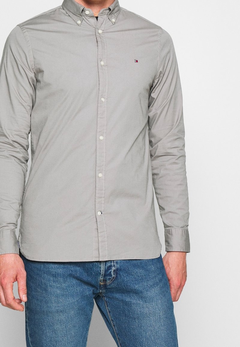 Homme portant une chemise gris clair à boutons manches longues et un jean bleu sur un fond uni.