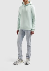 Vaaleanvihreä huppari pehmeästä kankaasta, jossa kengurun tasku ja nyörillinen huppu, yhdistetty vaaleansinisiin farkkuihin ja monivärisiin tennareihin.