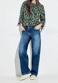 Donna che indossa una blusa a stampa verde scuro, jeans a palazzo blu con doppio bottone e sneakers beige chiare, in piedi con le mani nelle tasche.