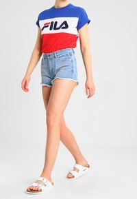 En person som bär en blå, vit och röd FILA-t-shirt, högmidjade fransade jeansshorts och vita sandaler med spänne, går framför en enfärgad bakgrund.