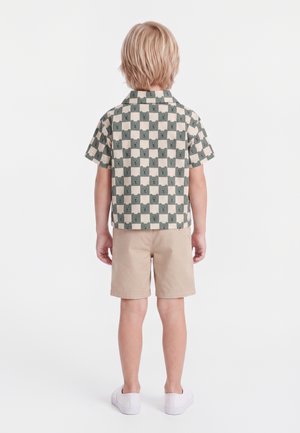 Enfant blond vu de dos portant un t-shirt à manches courtes avec des visages d’ours gris sur des carrés beige, un short beige et des chaussures blanches sans lacets.