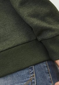 Sudadera verde oscura hecha de tela texturizada con puños y dobladillo acanalados, combinada con vaqueros de mezclilla azules que presentan costuras naranjas.