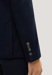 Marineblauwe blazer met een visgraatstructuur, voorzien van vier zwarte knopen aan de manchetten en een getailleerde pasvorm.