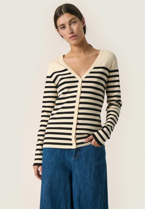 Stribet cardigan med dyb V-hals, beige og sorte striber, knaplukning foran, lange ærmer og ribbede manchetter, parret med brede denimbukser.