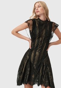 Donna in un abito di pizzo nero e oro con maniche corte trasparenti, in piedi con le mani sui fianchi, che guarda verso sinistra su uno sfondo neutro.