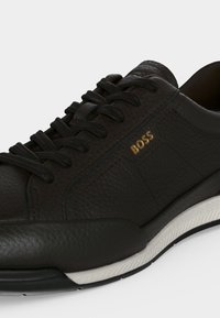 Baskets en cuir noir à surface texturée, lacets noirs, semelle blanche et noire, et logo doré "BOSS" sur le côté près des lacets.