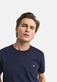 Camiseta de algodón azul marino con cuello redondo, que presenta un pequeño logo en el lado izquierdo del pecho. La tela parece suave y lisa.