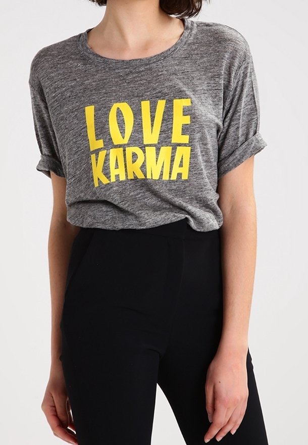 Personne portant un t-shirt gris à manches retroussées affichant en grand texte jaune "LOVE KARMA", rentré dans un pantalon noir taille haute.