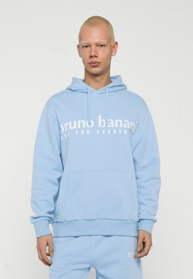 Personne aux cheveux blonds courts portant un sweat à capuche bleu clair et un pantalon assorti avec le texte blanc « bruno banani » sur le sweat.