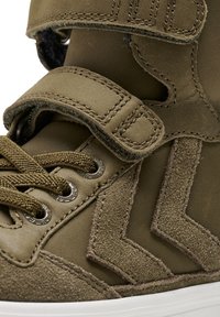 Zielone zamszowe sneakersy typu high-top z bocznym wzorem w kształcie chevron, skórzanymi paskami, metalowymi oczkami i teksturowanymi sznurowadłami. Biała gumowa podeszwa widoczna poniżej.