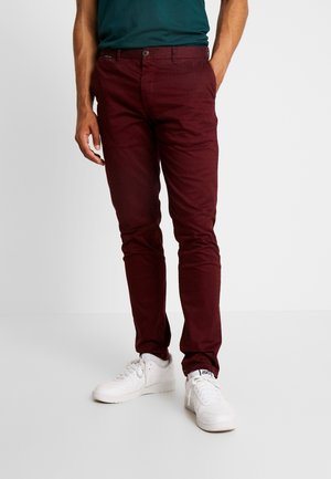 Homme portant un pantalon bordeaux ajusté, une chemise vert foncé, et des baskets blanches, debout devant un fond uni.