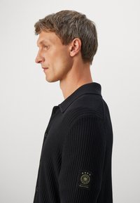 Perfil de un hombre con cabello corto que lleva un pulóver negro de canalé con un parche de logo en la parte superior de la manga izquierda.