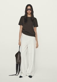 Brązowa koszulka z krótkim rękawem w połączeniu z białymi spodniami o szerokich nogawkach. Modelka trzyma ciemnobrązową torbę na pasku. Ciemne okulary dopełniają całość.