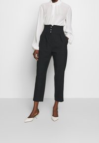 Högmidjade svarta byxor med veck, med framknappar och avsmalnande ben. Kombineras med en vit långärmad blus och beige klackar.