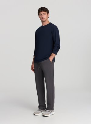 Giovane uomo in piedi, che indossa una maglia a maniche lunghe blu navy, pantaloni grigio scuro e sneakers di colore chiaro, con una mano in tasca.