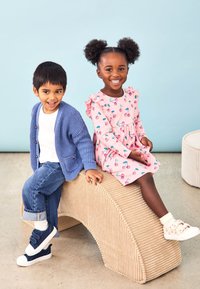 Cardigan tricotat albastru, tricou alb și blugi pe un copil; rochie roz cu imprimeu de cireșe și mâneci cu volane pe celălalt; bancă texturată bej.