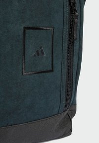 Dark teal fabric backpack featuring a black zip, square logo patch, and reinforced black base. Smooth texture with subtle pattern detailing.