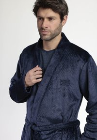 Hombre vistiendo una bata de felpa azul oscuro texturizada, atada a la cintura, sosteniendo el cuello con una mano, contra un fondo blanco liso.