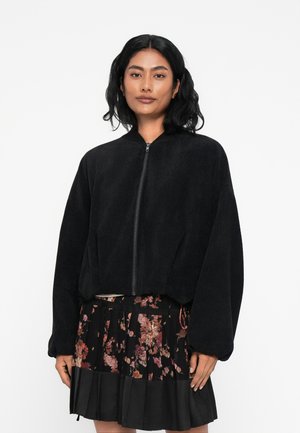 Giacca bomber in velluto a coste nera con chiusura a zip e polsini a coste, caratterizzata da un design cropped. Indossata sopra una gonna plissè floreale.