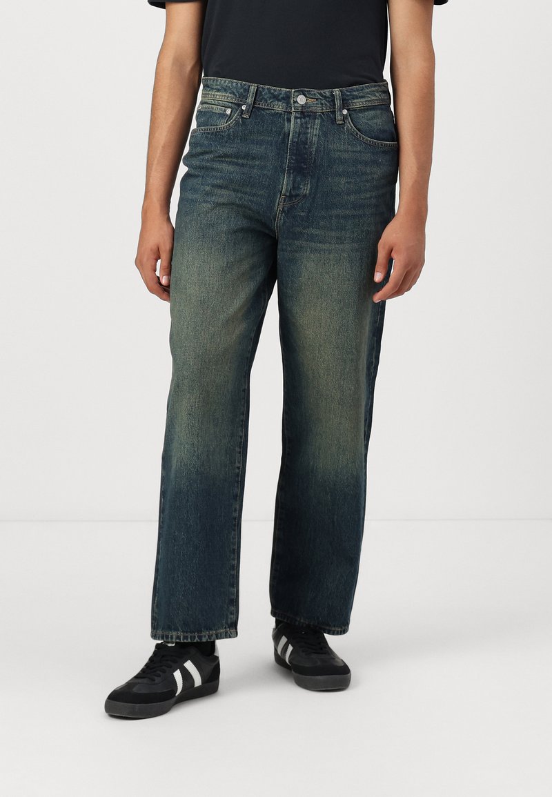 Jean en denim bleu avec une coupe droite, un léger délavage et un design à cinq poches. Portés avec des chaussures noires dotées de rayures blanches.