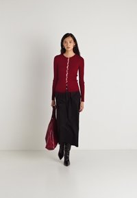 Raudona ilga rankovė kardiganas su mygtukais, derinamas su juoda maksi sijonu ir juodomis iki kelių botomis, laikantis bordo spalvos dideliu maišu. Lygi tekstūra.