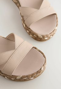 Sandales beiges avec des brides croisées en cuir texturé et des plateformes en corde tissée ornées de fils dorés sur un fond blanc.