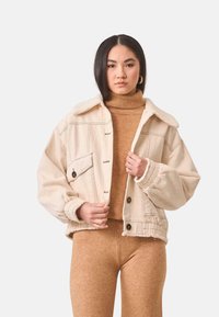 Chaqueta de mezclilla beige recortada con cuello de sherpa, grandes botones y puños elásticos, combinada con un cuello de tortuga marrón a juego y leggings.