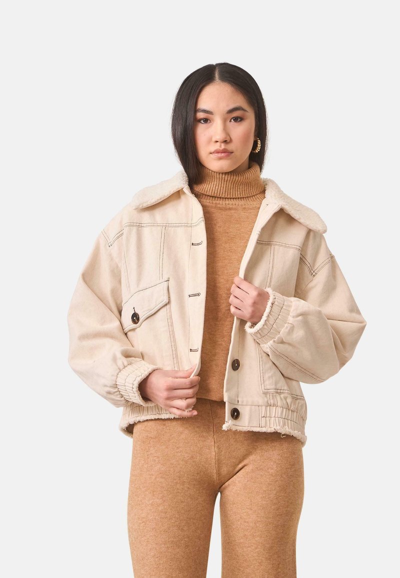 Chaqueta de mezclilla beige recortada con cuello de sherpa, grandes botones y puños elásticos, combinada con un cuello de tortuga marrón a juego y leggings.