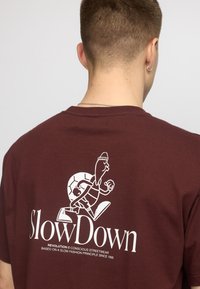 Bordeaux katoenen t-shirt met een witte afbeelding van een karakter en de tekst "SlowDown" op de achterkant. Ronde hals met korte mouwen.