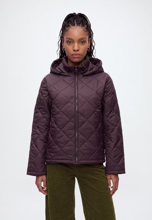 Femme aux cheveux tressés portant une veste matelassée zippée violet foncé avec capuche et un pantalon vert olive, debout devant un fond clair.
