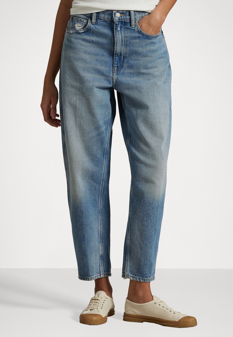 Pessoa a usar jeans de perna reta em azul claro desbotado, com a mão no bolso, e tênis de lona bege com solas castanhas.