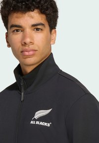 Sort jakke med høj krave, lynlås foran og broderet hvidt bregne-logo. Lavet af glat, strækbart materiale med subtile skulderdetaljer.