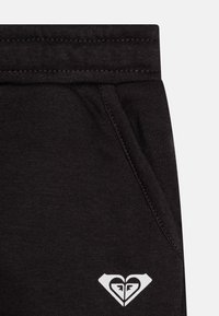 Pantalons de survêtement en coton noir avec une poche latérale, une taille élastique et un logo blanc en forme de cœur en bas à droite.