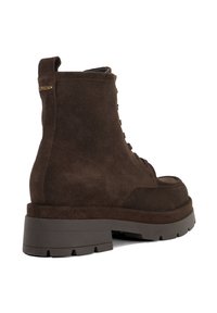 Brown suede ankle boot with a thick rubber sole, lace-up design, and a pull tab at the back; gold branding detail near the top.