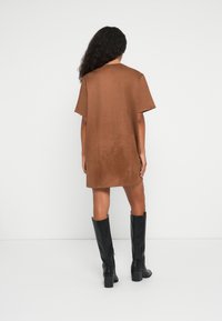 Vrouw met lang krullend haar, gekleed in een losse bruine jurk met korte mouwen en zwart leren laarzen tot de knie, staande tegen een witte muur.