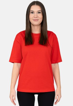 Young woman with long brown hair wearing a loose red short-sleeve t-shirt and black pants, standing against a plain white background.