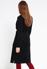 Abito avvolgente nero a maniche lunghe in tessuto morbido e strutturato con cintura in vita; presenta una silhouette fluida e spacchi laterali, indossato sopra collant rossi.